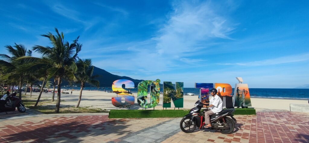 Vietnamese man riding a motorbike along Da Nang beach with sea and mountains in the background – a typical scene along Vietnam’s coastal travel routes